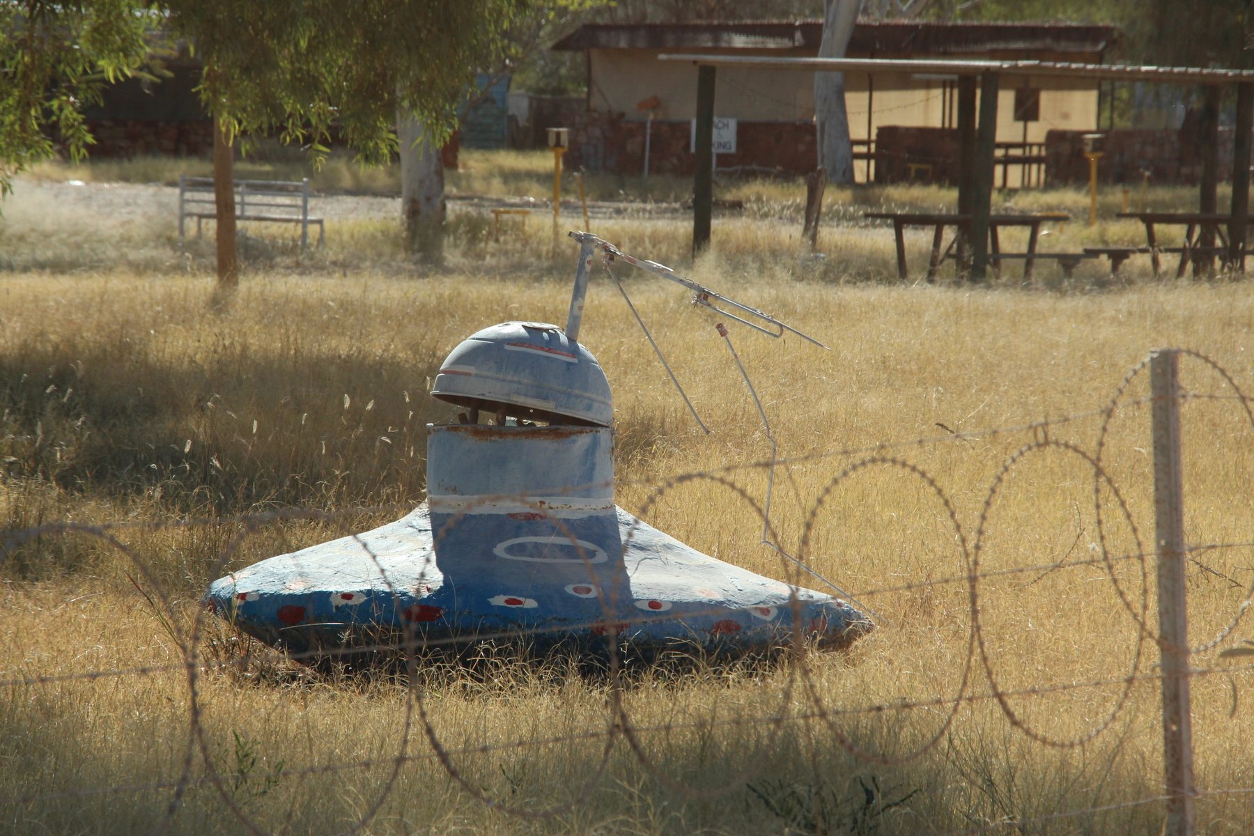 A weathered, alien-themed UFO sculpture standing in overgrown grass behind razor wire at the abandoned Wycliffe Well roadhouse, Northern Territory, Australia